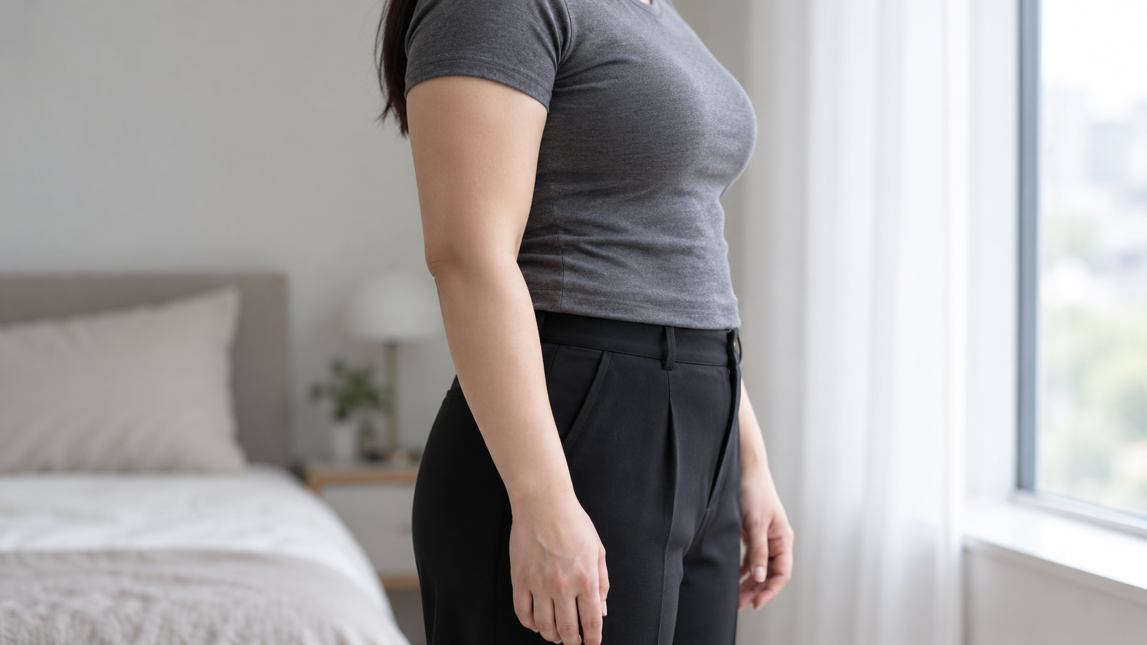 짧은 소매 회색 상의를 입은 여성이 창가에서 옆으로 서 있는 모습